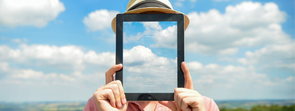 Man with Ipad covering his face which a screensaver which matches the sky and clouds in the background giving the illusion of transparency.